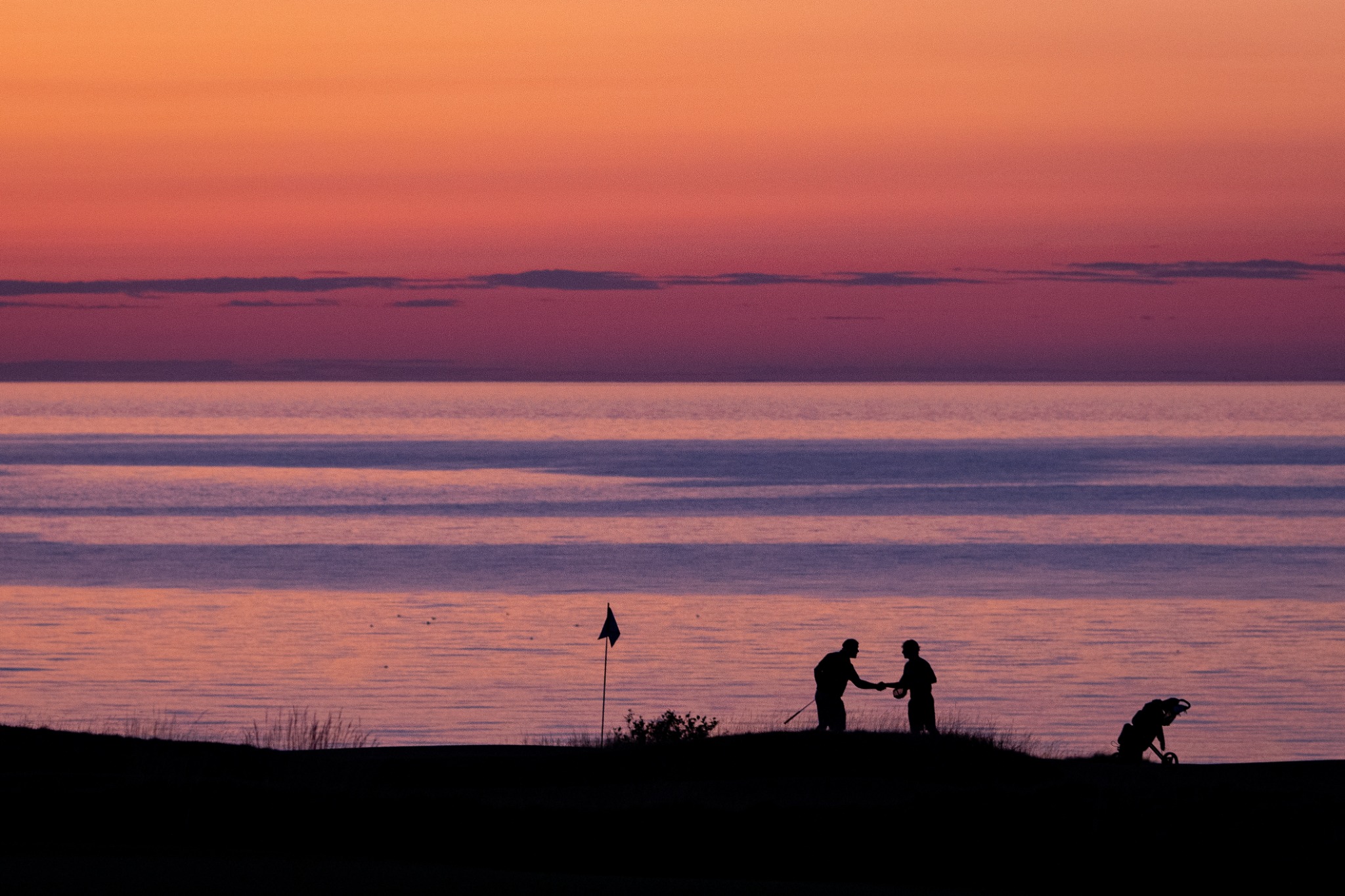 Two golfers shake hands beside the ocean as an orange and pink sunset settles over the course